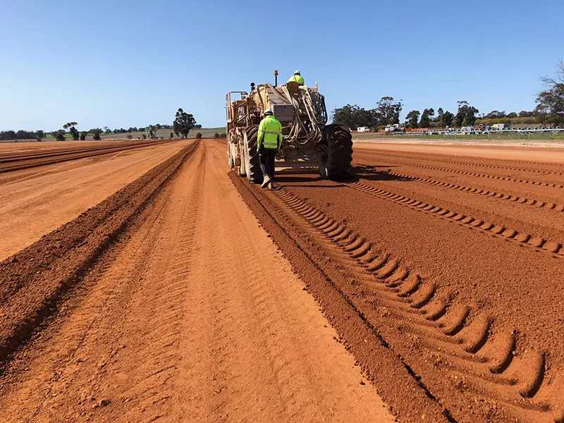 Workers operate a large machine on a red dirt agricultural field under a clear blue sky, creating evenly spaced rows.