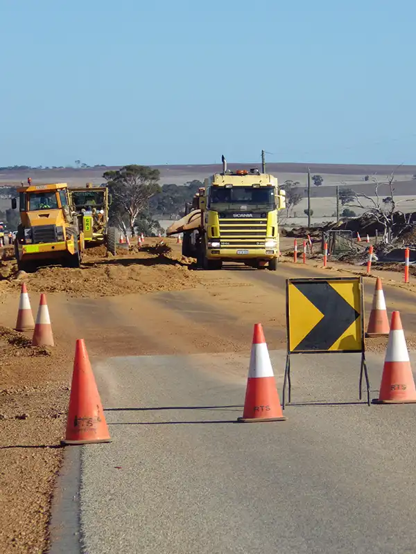 A road construction scene with trucks and equipment, orange cones, and a right-turn warning sign on a rural road showcases vital work, including soil stabilisation efforts.