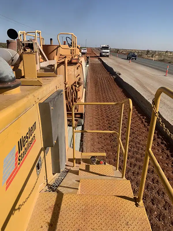 A road stabilization machine with a "Western Stabilisers" logo is performing soil stabilisation on a dirt road next to a paved road with traffic.