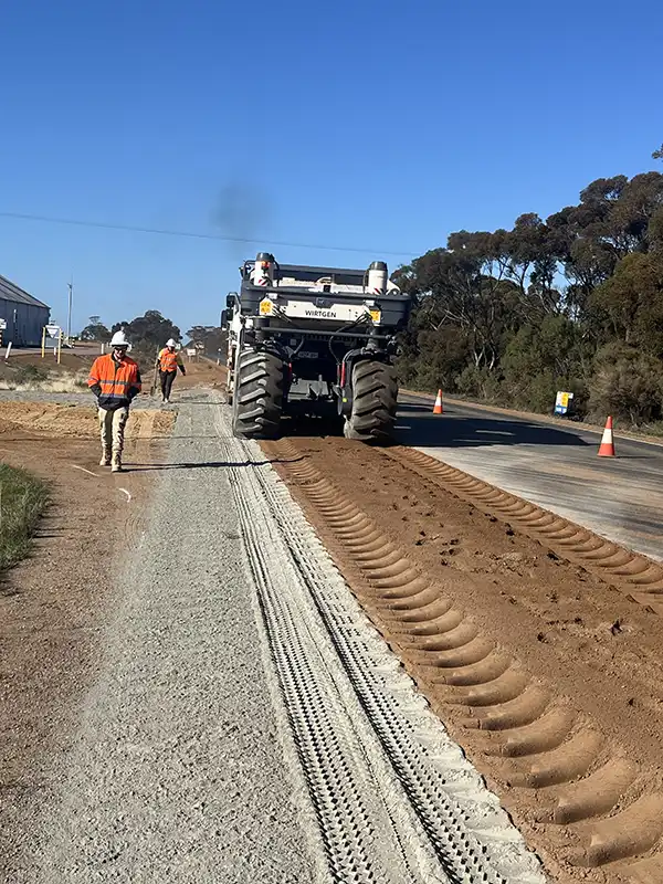 Workers in orange jackets operate a large road construction machine, which is laying fresh dirt on a rural road as part of essential soil stabilisation efforts.