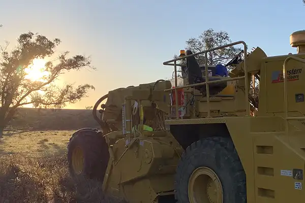 A large yellow mining vehicle in use at sunrise, with a tree on the left and dusty terrain in the background, showcases early morning soil stabilisation efforts.