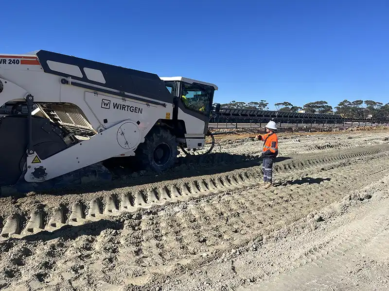 Construction worker in an orange and blue uniform operates a Wirtgen machine on a large, dirt construction site under a clear blue sky.