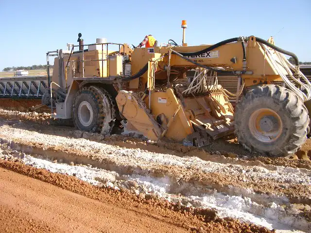 A large, yellow construction machine with large tires is operating on a dirt site, surrounded by a barren landscape under a clear sky, working efficiently on soil stabilisation.