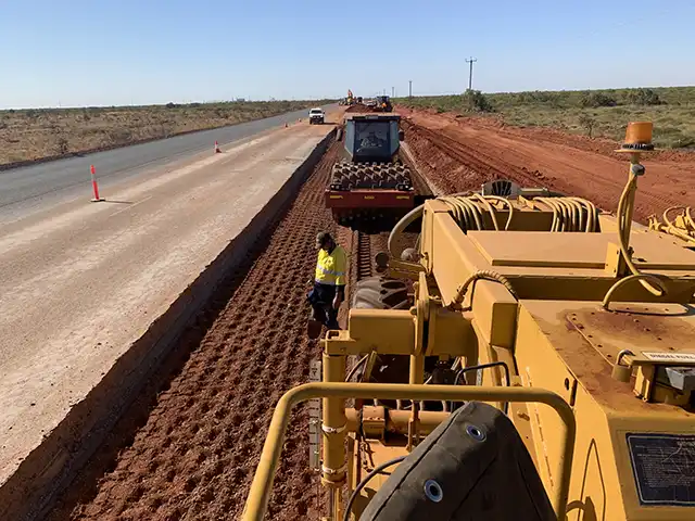 Construction workers using heavy machinery are busy on a dirt road, engaging in soil stabilisation efforts; a new paved road is under construction nearby.