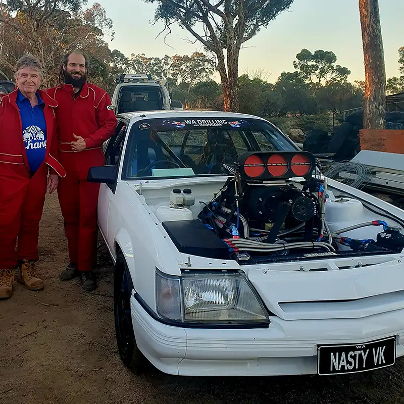 Two people in red suits standing beside a white drag race car with an exposed engine. The car has the license plate "NASTY VK".