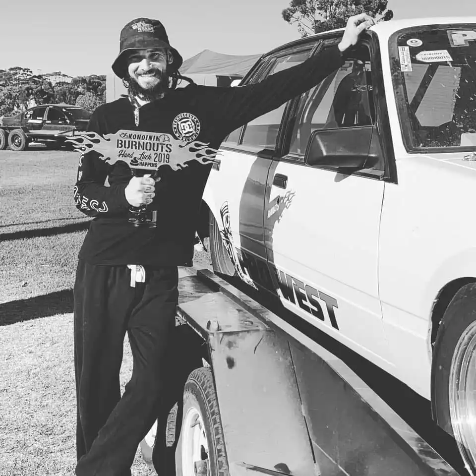 A person in a hat and sunglasses leans on a car while holding a 2019 Championship Burnouts award.