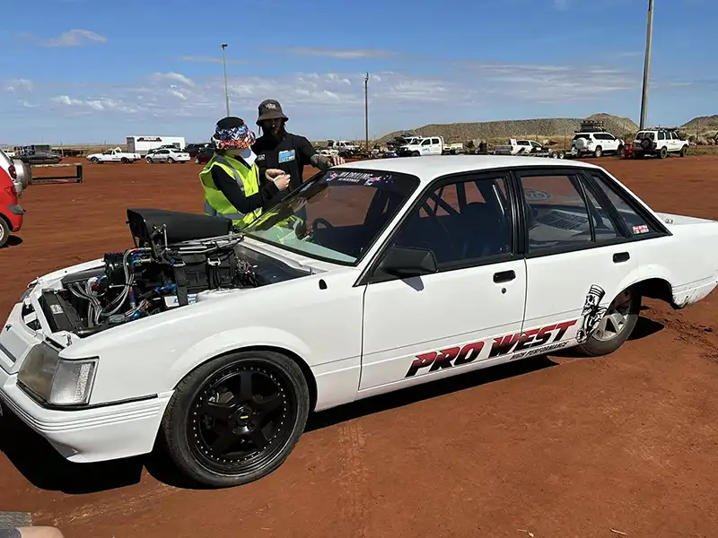 A white car with “Pro West” written on the side is parked on a dusty ground with two people standing next to it.