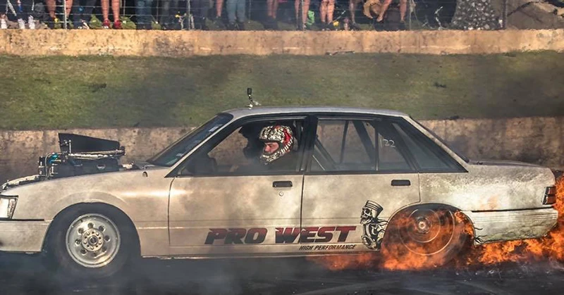 A vehicle marked "Pro West High Performance" with rear tires on fire, driven in a motorsport event while spectators watch.