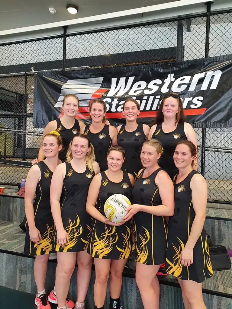 A women's netball team poses for a photo in matching black and yellow uniforms, with a banner reading "Western Stallions" in the background.