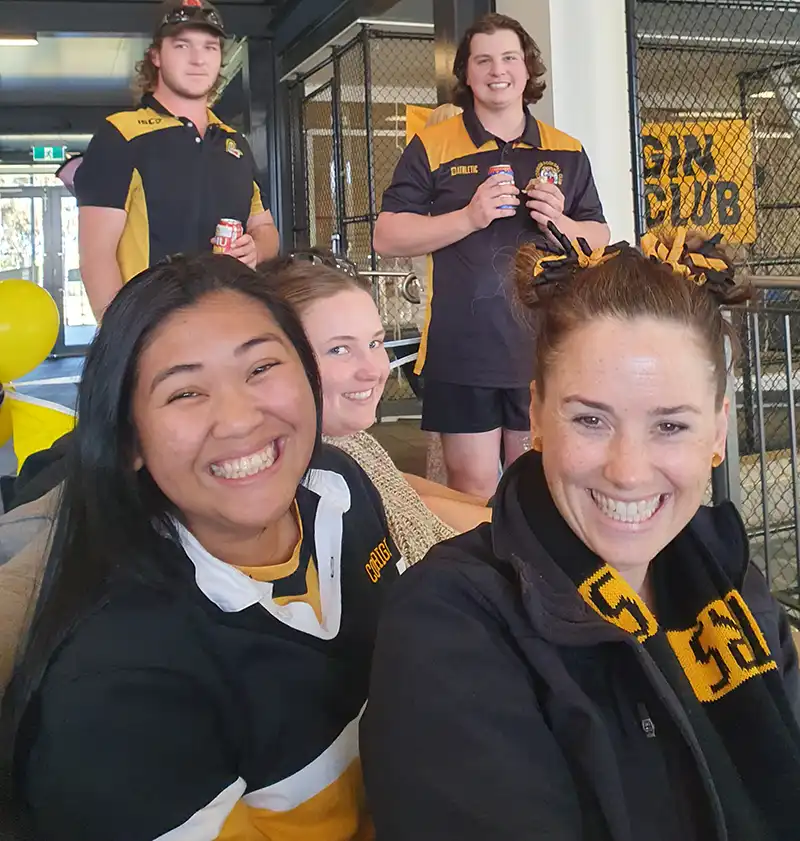 A group of people wearing black and yellow sports jerseys and scarves smiling indoors. Yellow balloons are visible in the background.