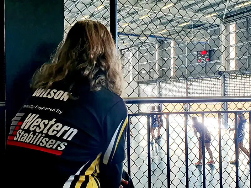 A person in a sports jersey watches a group playing netball inside a caged indoor court.