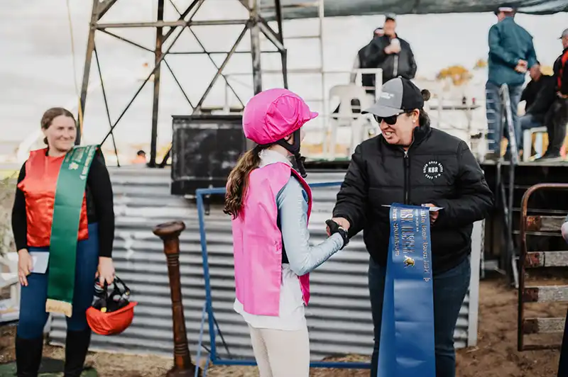 A person in a pink jockey outfit receives a blue ribbon from another person in a cap at an outdoor event.