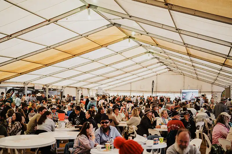 Crowd seated at round tables inside a large event tent, engaged in conversations, eating, and enjoying the lively atmosphere.