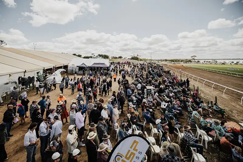 Crowd gathers at an outdoor horse racing event near a track, with tents and seating areas under a partly cloudy sky.