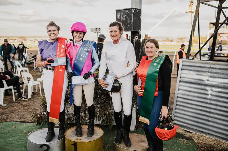 Four equestrians in riding gear stand on podiums, holding ribbons and envelopes, with a racetrack in the background.