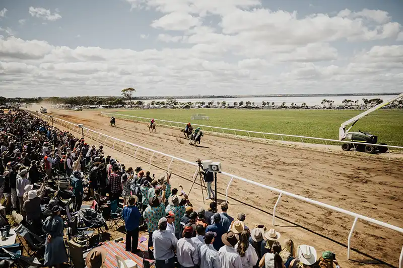 Horse race on a dirt track with spectators cheering on the sidelines. Sunny day with scattered clouds in the sky.