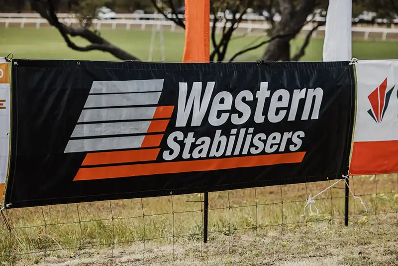 Banner with "Western Stabilisers" logo and text on a metal fence, set in a grassy outdoor area with trees in the background.