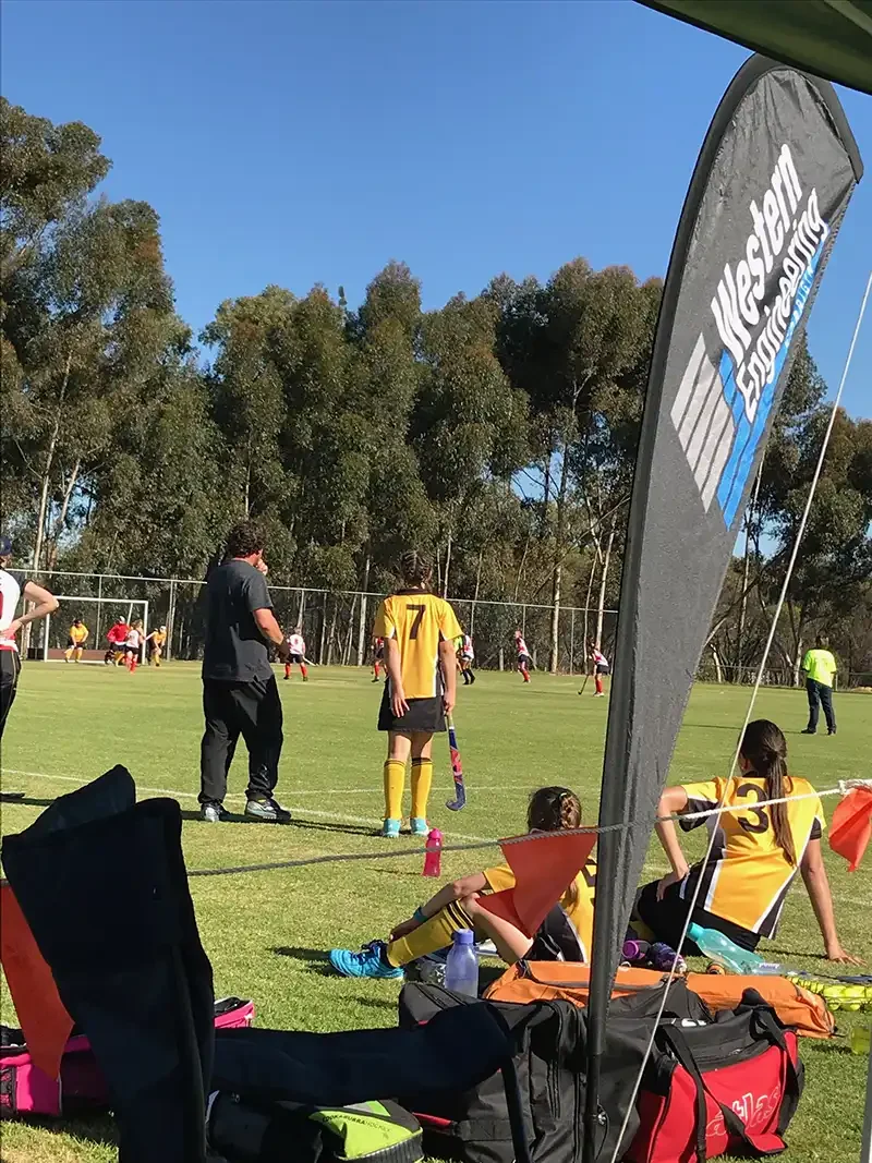 People watching a field hockey game with players in uniform on a grassy field; a banner and trees in the background.